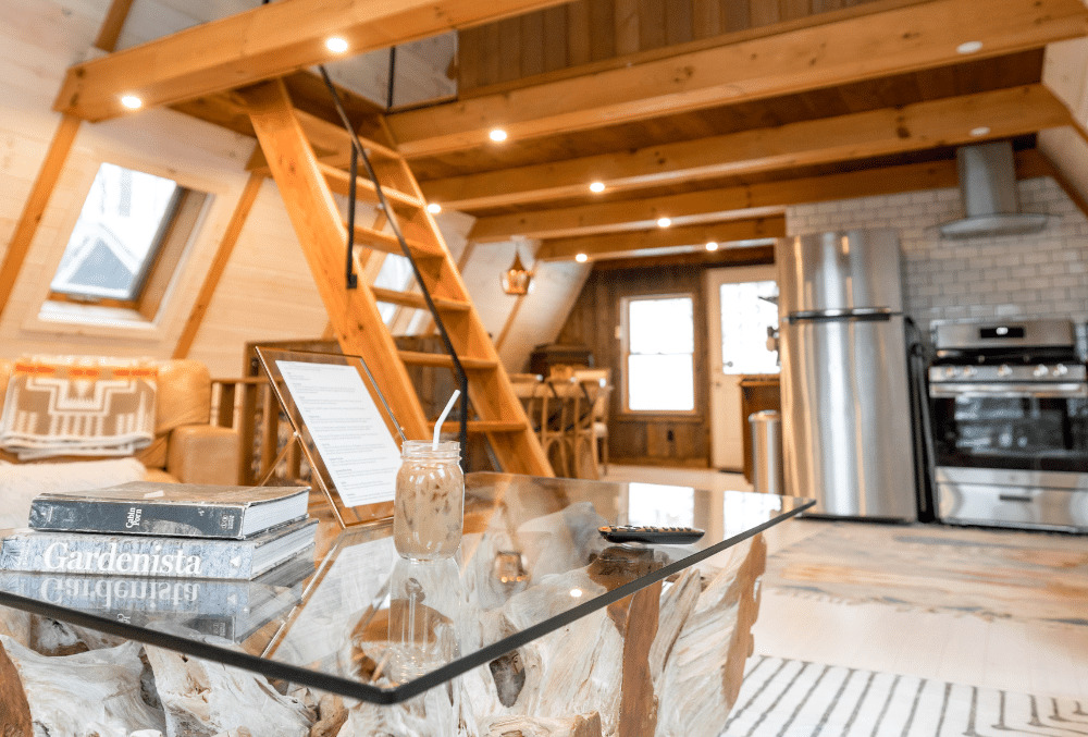 Cozy and well-lit cabin interior with a kitchenette, loft ladder, and wooden furnishings, featuring a marble coffee table with books and an iced drink in the foreground.