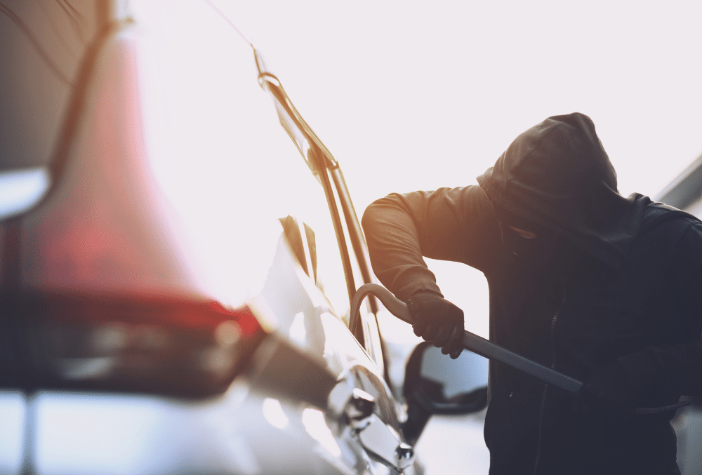 Silhouette of a person in a hooded jacket using a crowbar to break into a car.