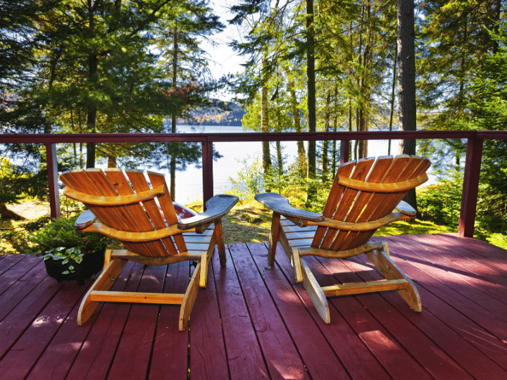 Two wooden Adirondack chairs on a red deck overlooking a tranquil lake surrounded by evergreen trees.