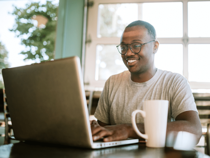Smiling entrepreneur working on a laptop with a coffee mug on the table in a bright office space.