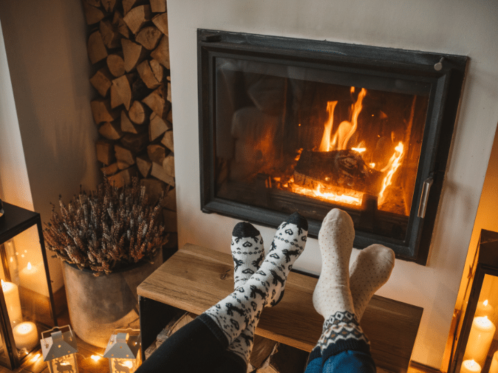 Two people with their feet up near a cozy fireplace, surrounded by candles and a woodpile.