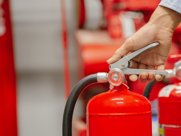 A hand performing maintenance on a red fire extinguisher with a pressure gauge, against a background of multiple extinguishers.