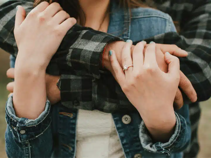 Close-up of a woman's hand with an engagement ring, embraced by a person in a plaid shirt.