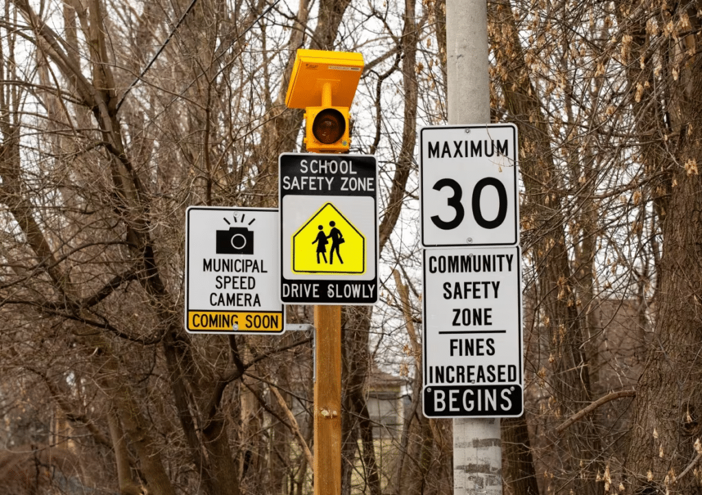 A collection of traffic signs including a 'Municipal Speed Camera Coming Soon' notice, a 'School Safety Zone' sign, and a 'Maximum 30 - Community Safety Zone Fines Increased Begins' sign.