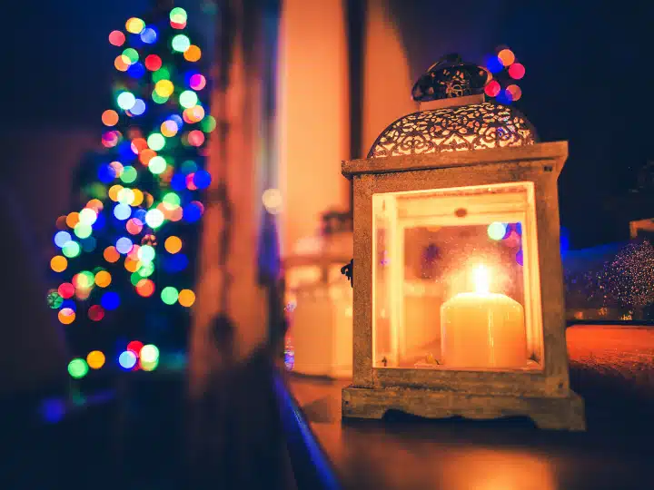 A candle inside a decorative lantern with a blurred background of a Christmas tree adorned with colorful lights.