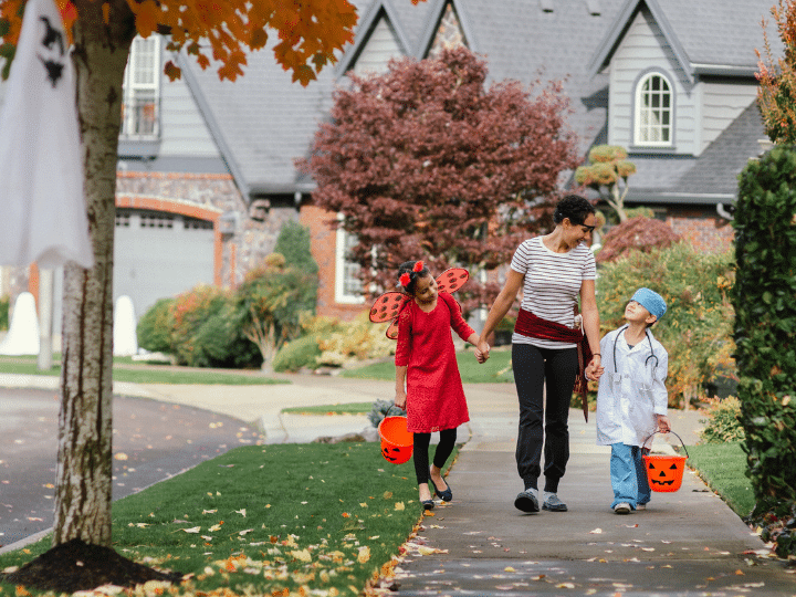 A parent holding hands with two children dressed in Halloween costumes, walking on a sidewalk lined with autumn leaves.