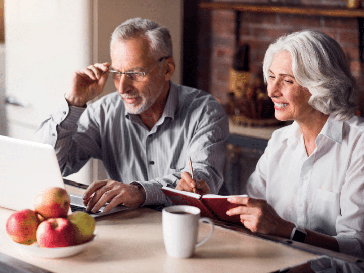 older couple sitting side-by-side at their kitchen table, looking intently at a laptop and studying what's on it's screen.