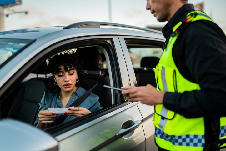A female driver with curly hair looking at a document while being pulled over by a police officer, illustrating the consequences of breaking G2 driving restrictions.