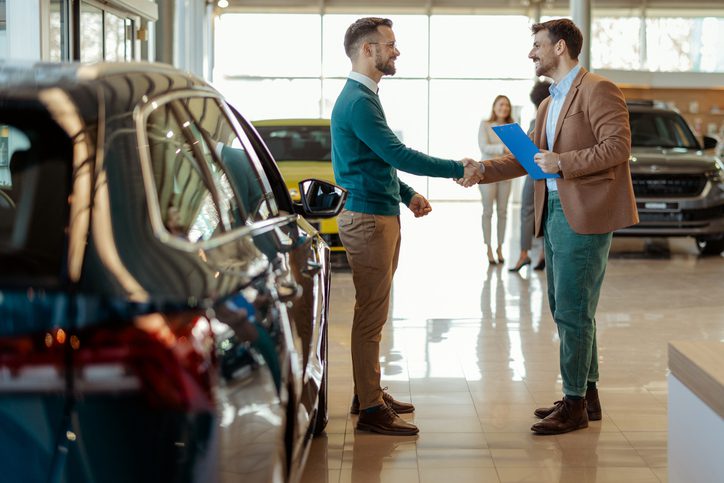 Two men shaking hands in a car dealership next to a new car, discussing how using Anti-Theft Devices Matter for Insurance rates.