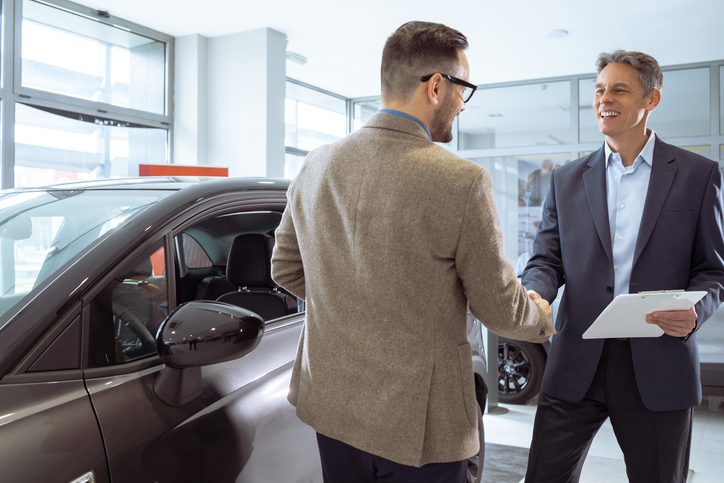 Two men, one customer and one salesman, shaking hands inside a car dealership next to a new vehicle, discussing potential liability for Theft Surcharge Insurance.