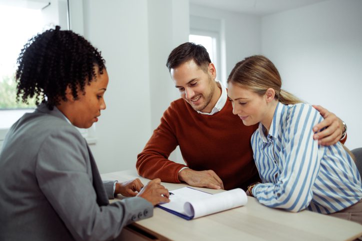 A professional broker explains the details of a policy document to a couple in a modern office, highlighting the broker’s role during a claim in ensuring clients understand their coverage.