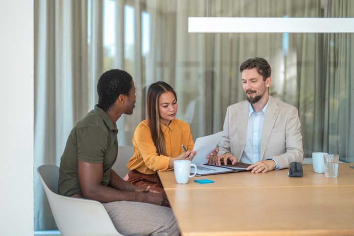 A professional insurance broker explains policy options to a couple during a meeting in a bright, modern office as the woman reviews a document, illustrating what insurance brokers do for clients.