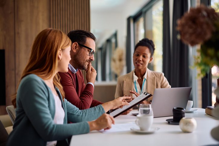 An insurance broker reviews a policy document with a couple at their home, explaining the terms on a clipboard while the man and woman look on thoughtfully over coffee.